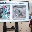 Two students, one dressed in black and one dressed in purple talking in front of an image of medical devices on a screen
